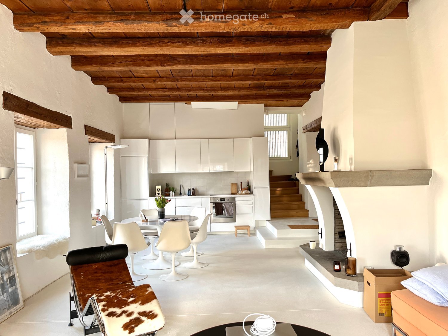 Open-plan living and dining area with a modern kitchen, white dining chairs, a leather sofa, and a fireplace. The space features exposed wooden beams and white walls.