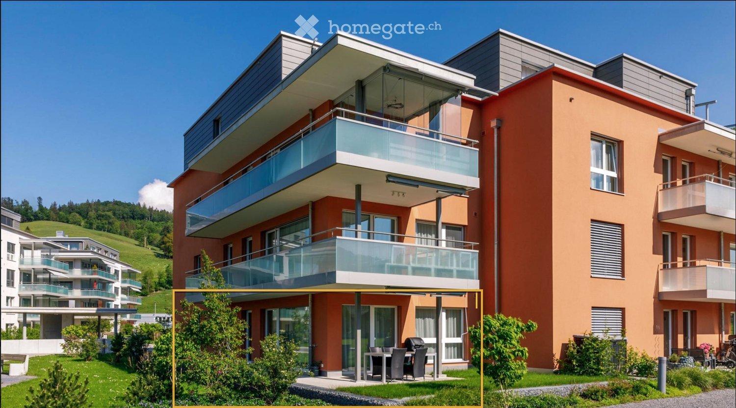 Modern multi-story residential building with orange facade, large windows, and balconies. The building is surrounded by greenery and has a well-maintained lawn and patio area with outdoor furniture.