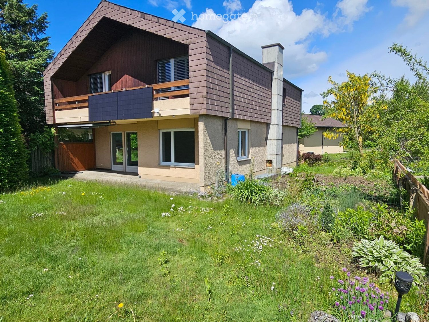 Two-story house with a modern design, featuring a balcony on the upper level, large windows, and a well-maintained garden with various plants and flowers.