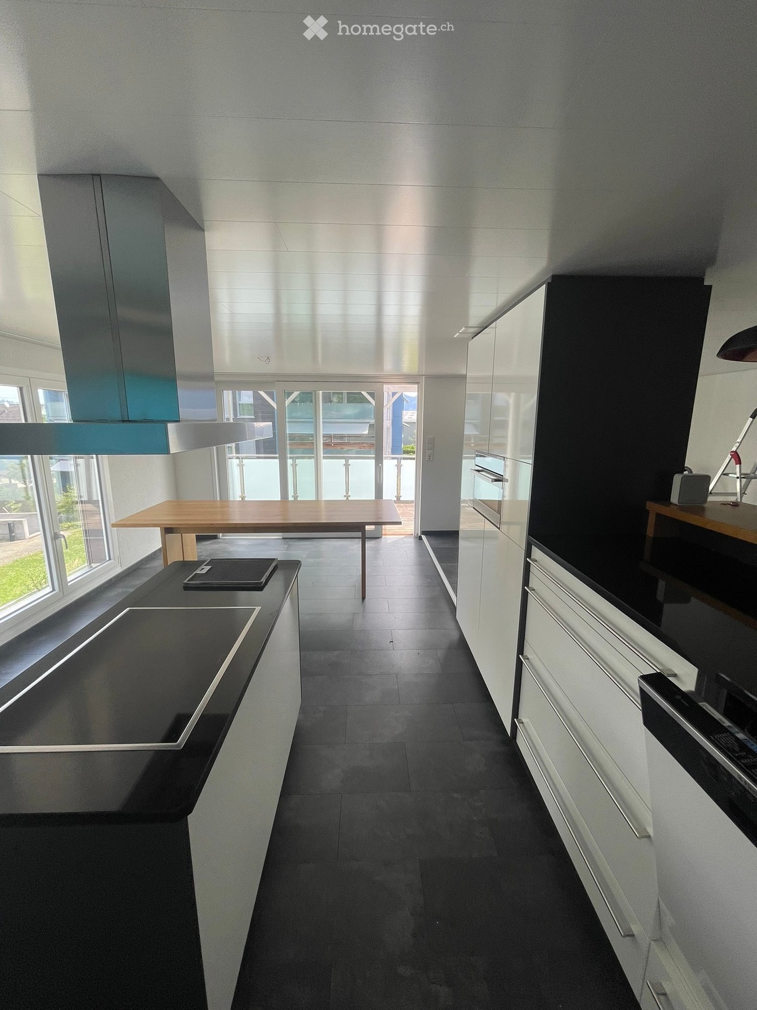 Modern kitchen with dark countertops, white cabinets, a wooden dining table, and a range hood. Large windows and a sliding glass door lead to an outdoor area.