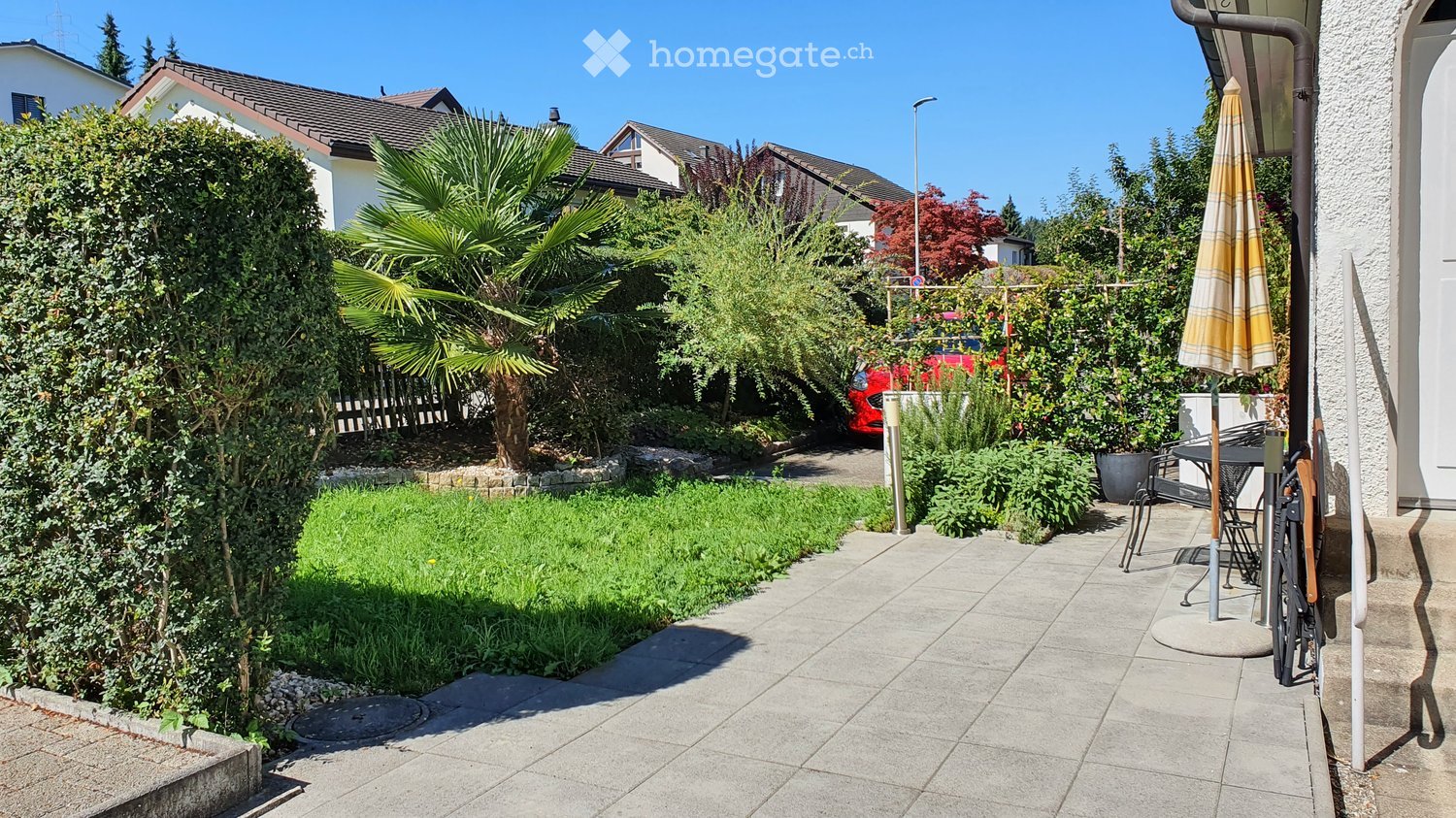 Well-maintained garden with a variety of plants, including a palm tree, shrubs, and a neatly trimmed lawn. A patio area with a table and chairs is visible, along with an umbrella and bicycles leaning against the wall.