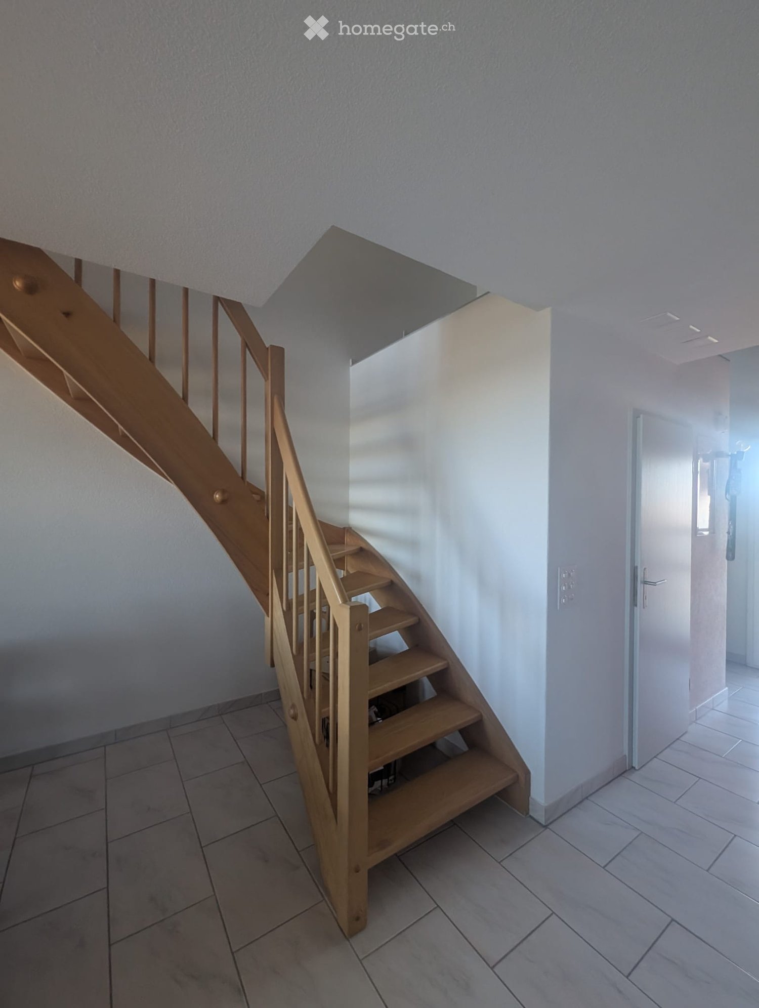 Wooden staircase with a handrail, leading to an upper level, with tiled floor and white walls.