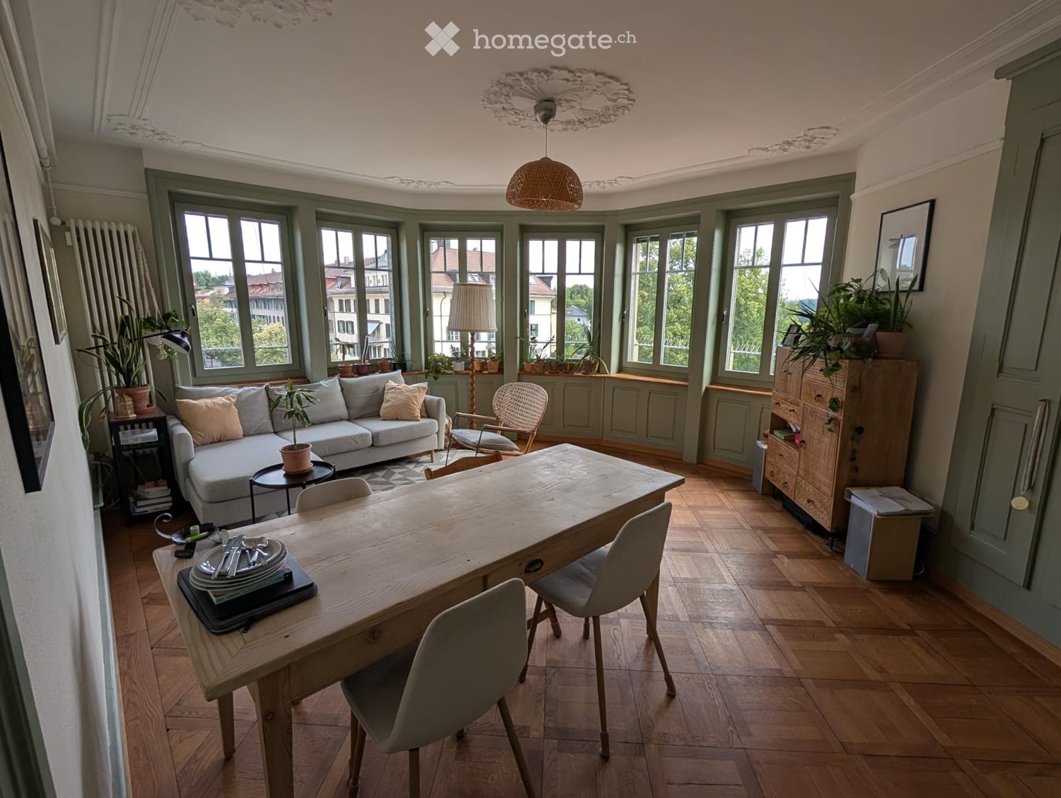 Bright living and dining area with large windows, wooden flooring, a grey sofa, a wooden dining table with chairs, and decorative plants.