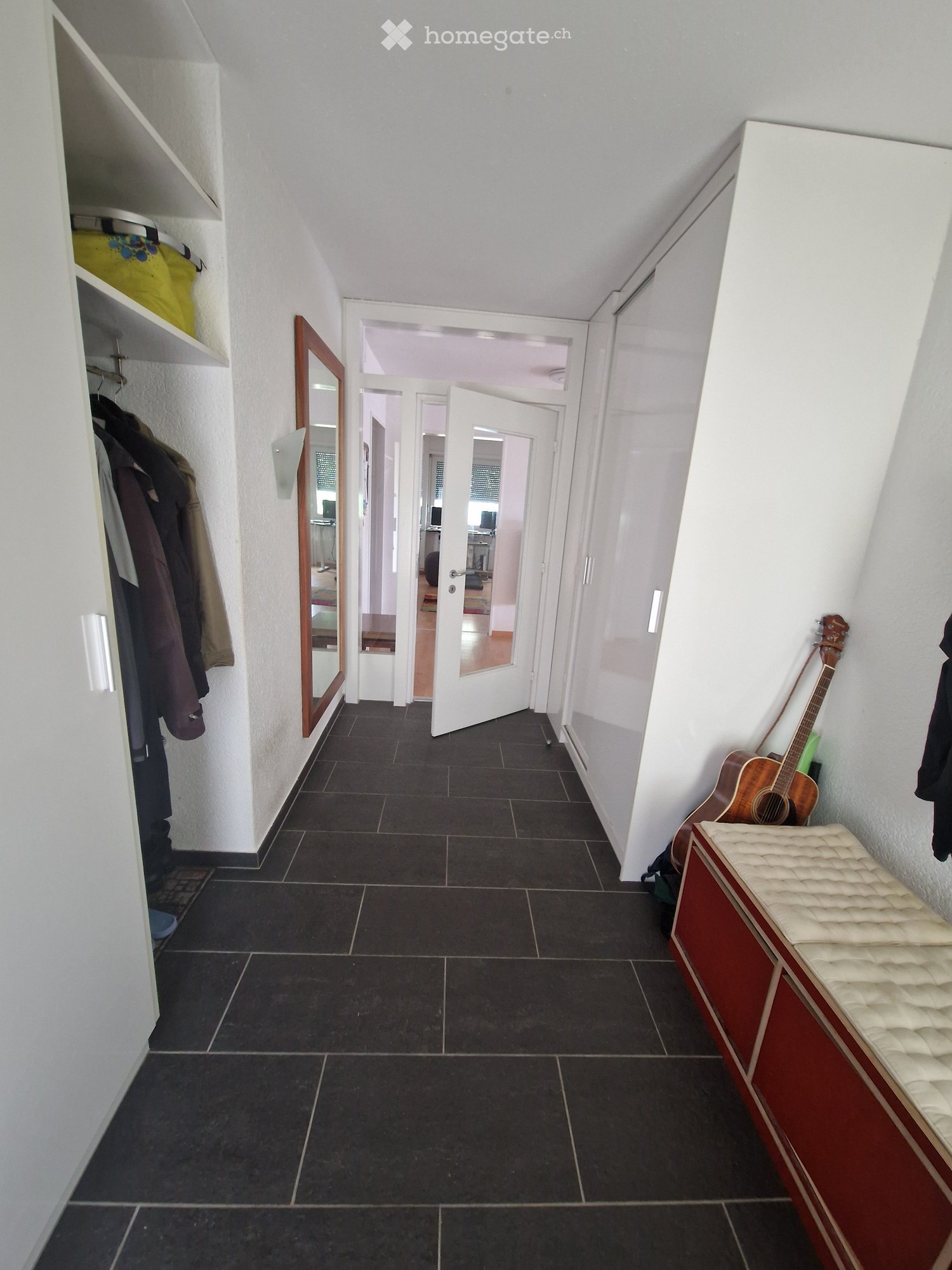A narrow hallway with dark tiled flooring, white walls, and several doors leading to other rooms. A mirror and coat rack are visible on the left, and a guitar and bench are on the right.