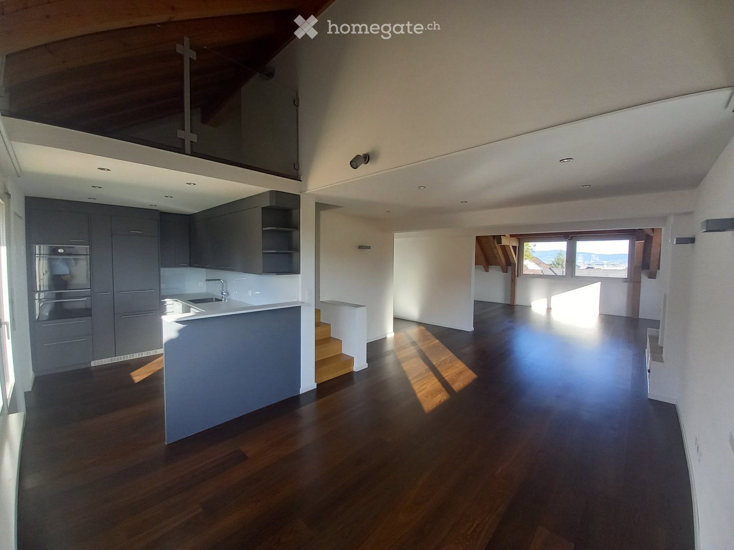 Open-plan space with a modern kitchen area featuring dark grey cabinets, a white countertop, and a sink. The main area has wooden flooring and large windows offering a scenic view. The ceiling is white with recessed lighting, and there is a small staircas