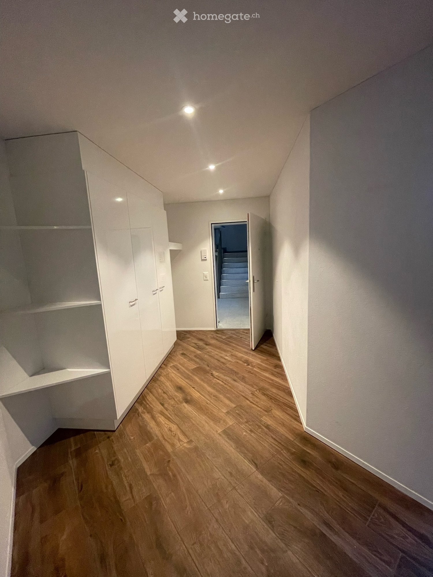 Empty hallway with wooden flooring, white walls, built-in storage unit on one side, and an open door leading to another room.