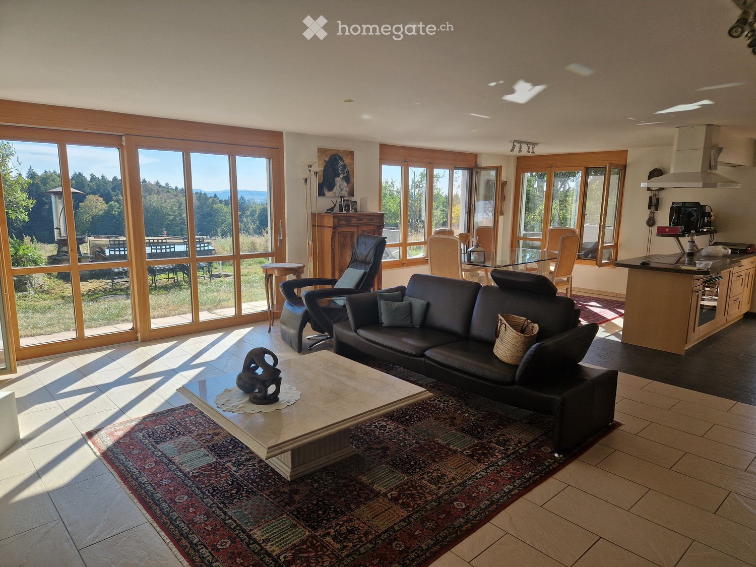 Open-concept living and dining area with large windows, a black leather sofa, a coffee table, a dining table with chairs, and a kitchen island with an oven.