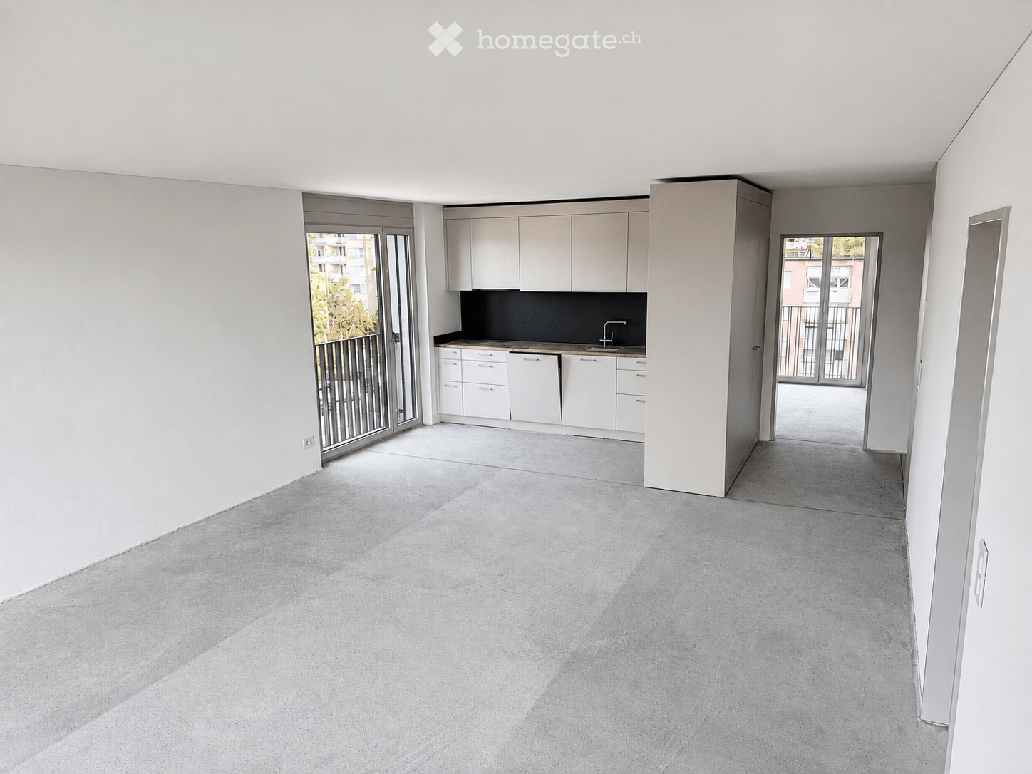 Empty room with white walls, grey concrete floor, and a small kitchen area with white cabinets and a sink.