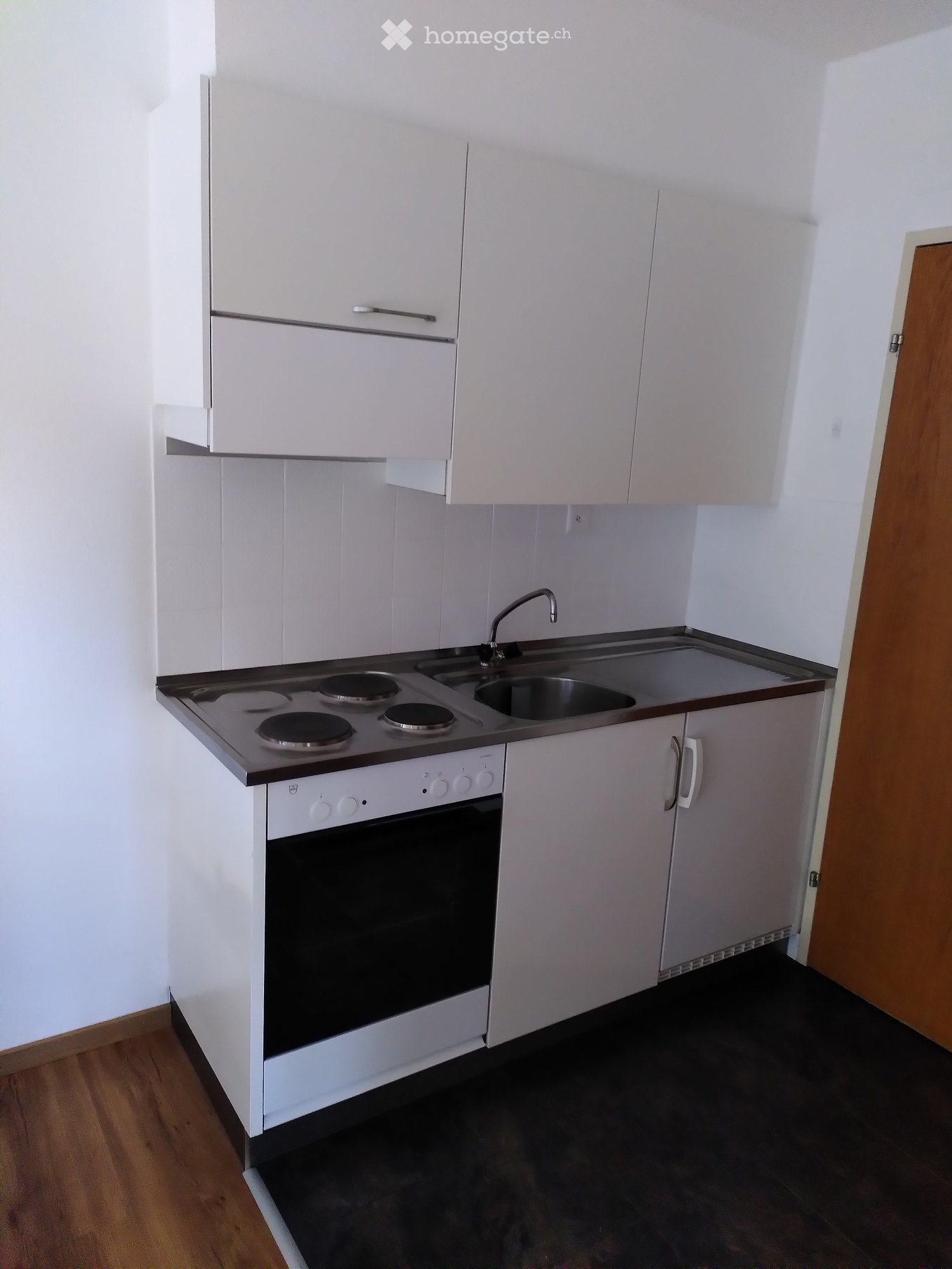 Modern kitchen with white cabinets, a four-burner electric stove, a sink with a faucet, and a built-in oven. The floor is dark, and the walls are white.