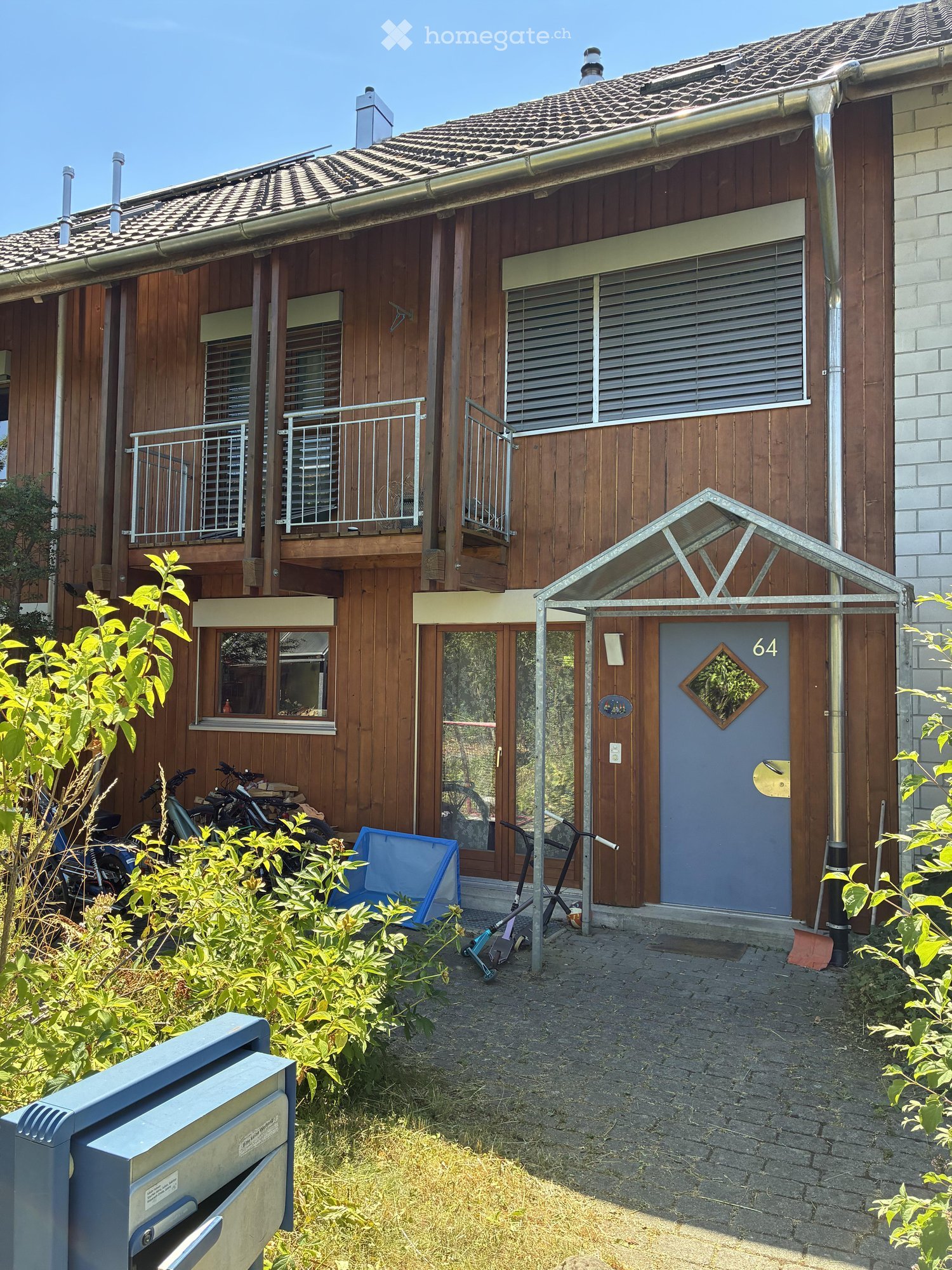 Two-story wooden house with a tiled roof, multiple windows, balconies, and a small entrance area with bicycles and a blue container.