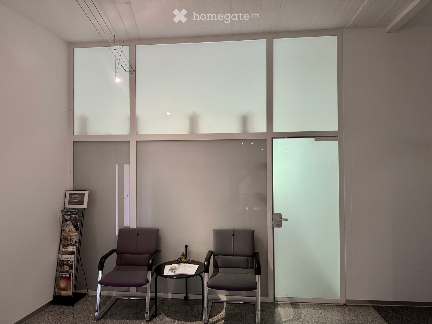 A small, modern waiting or reception area with two grey chairs, a small round table, a magazine rack, and a picture frame on the wall. Large windows and a frosted glass door are visible.