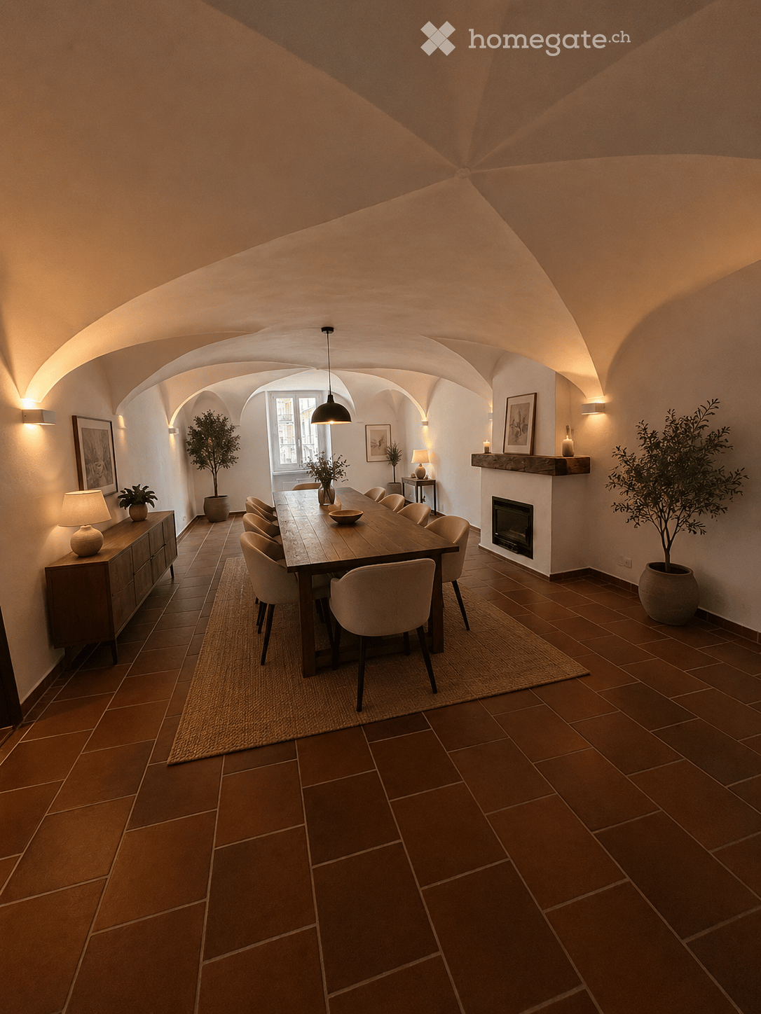 Elegant dining room with exposed wooden beams, terracotta tiles, a long wooden dining table surrounded by chairs, a fireplace, sideboard, table lamps, and potted plants.