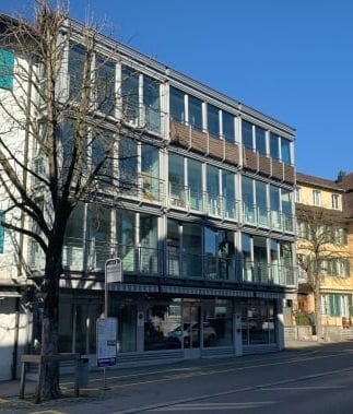 Modern multi-story building with large glass windows and balconies, located on a street with a clear blue sky.