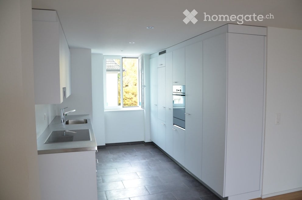 Modern, minimalist kitchen with white cabinets, stainless steel sink, and built-in oven. Features a large window with a view of greenery outside.