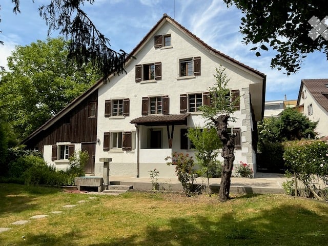 Two-story white house with brown shutters, wooden siding on the left side, and a small porch with a wooden awning. The house is surrounded by a well-maintained lawn and trees.