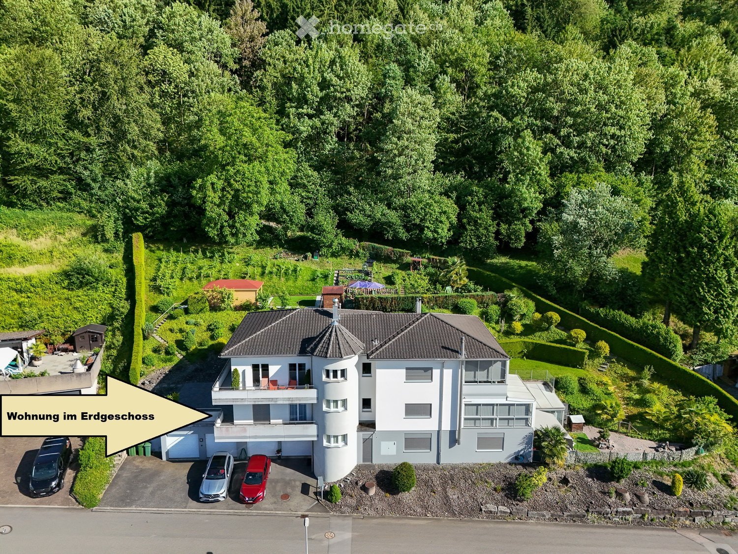 A modern two-story house with a raised ground floor, surrounded by lush greenery and trees. The house features a garage, multiple balconies, and a well-maintained garden area.