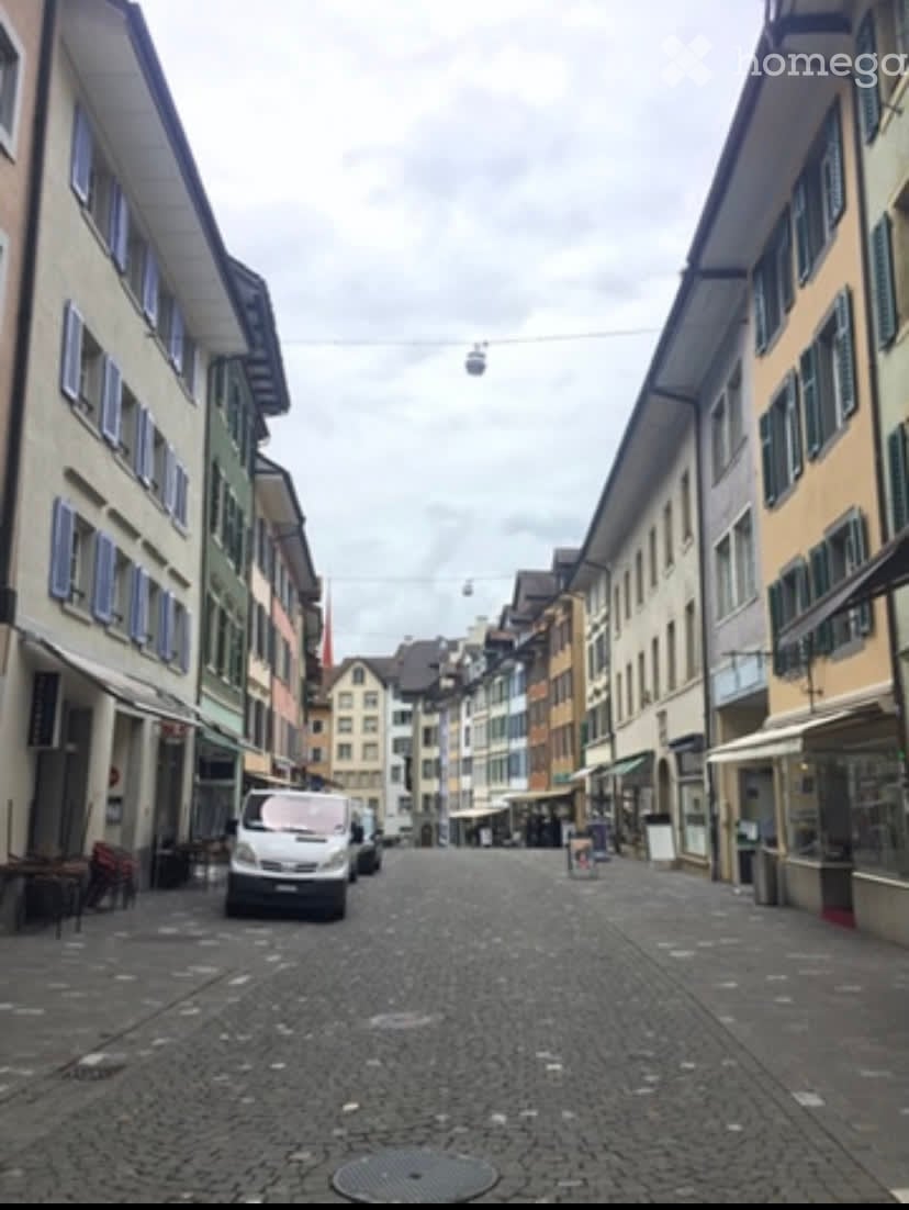 A quiet urban street lined with multi-story buildings, featuring colorful facades, shuttered windows, and a cable car system overhead.