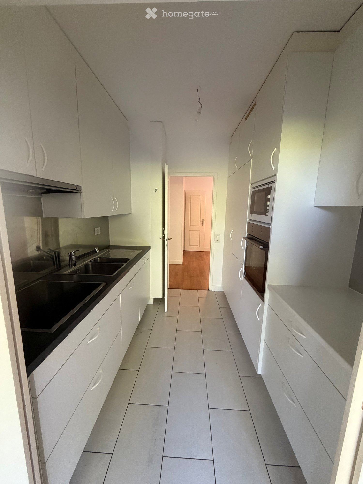 Modern kitchen with white cabinets, a double sink, microwave, and oven. The floor is tiled, and the walls are painted in a light color.