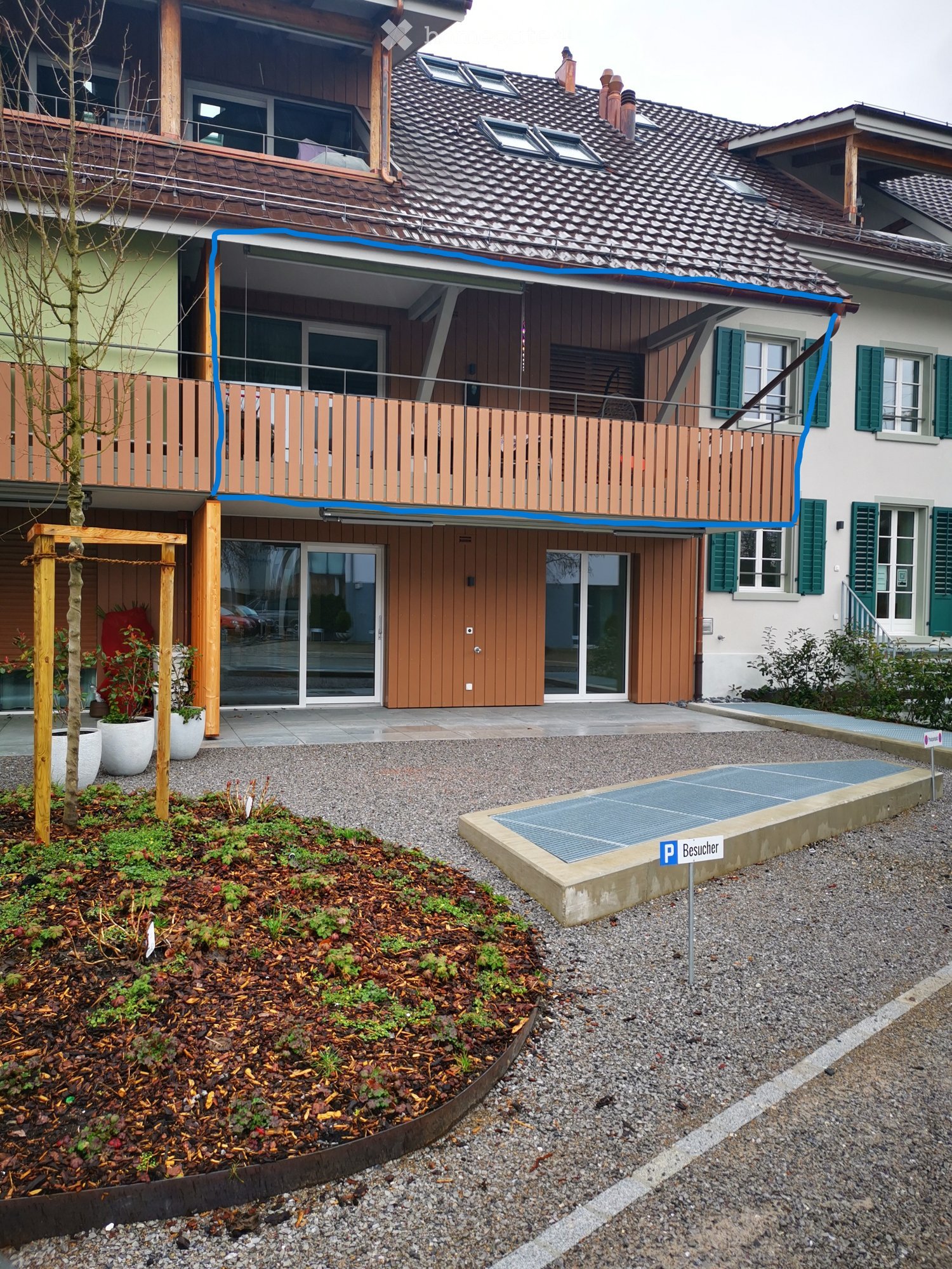 Two-story house with a balcony, wooden deck, and modern entrance. The house features a tiled roof, green shutters, and a small garden area with potted plants and a gravel driveway.