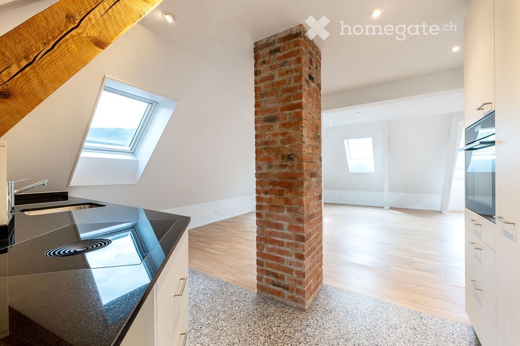 Open-plan attic space with exposed wooden beams, a brick pillar, and skylight windows. The area includes a modern kitchen counter with a sink and a built-in oven, but no furniture is present.