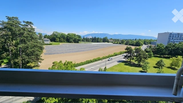 Balcony overlooking a landscape with a road, green fields, trees, and distant mountains under a clear blue sky.