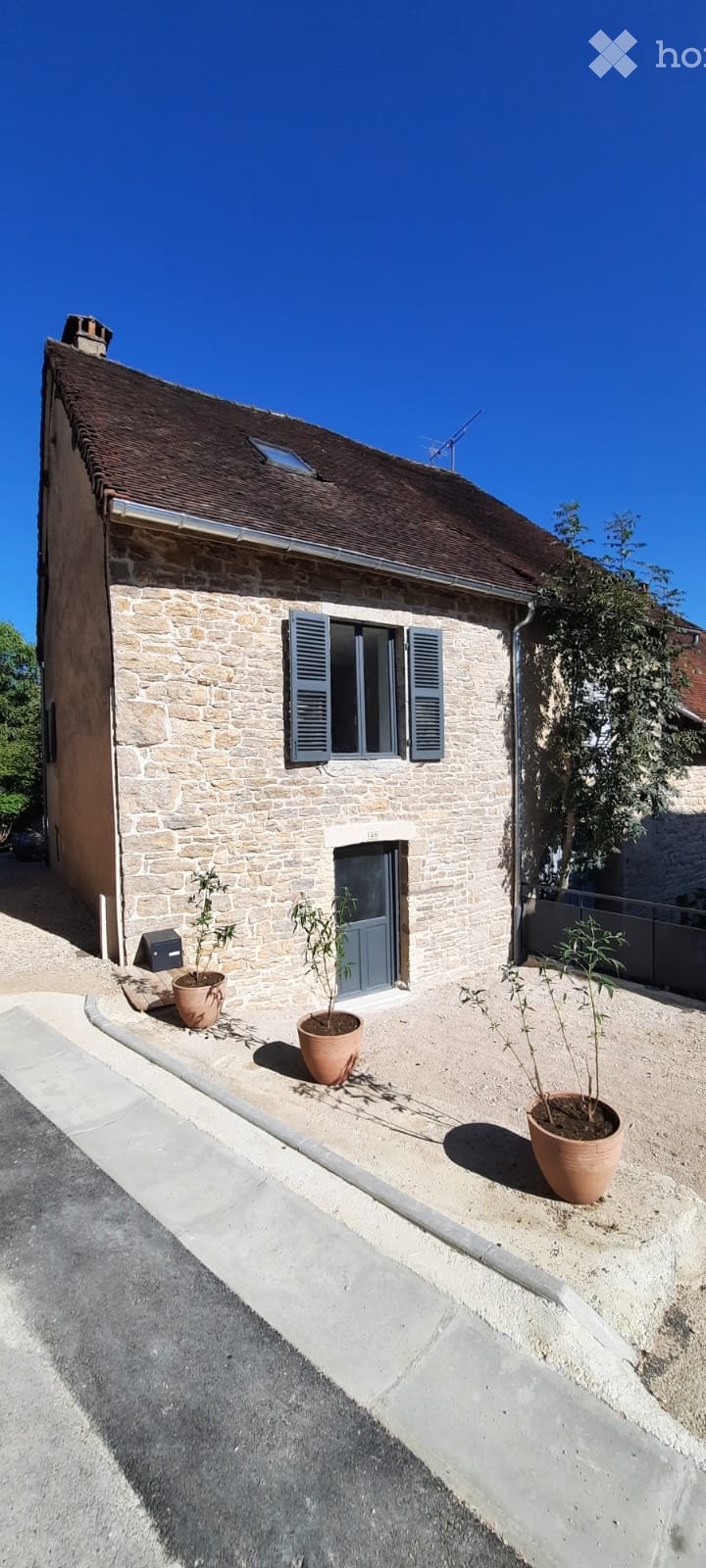 A stone house with a tiled roof, blue shutters, and a small garden area with potted plants.