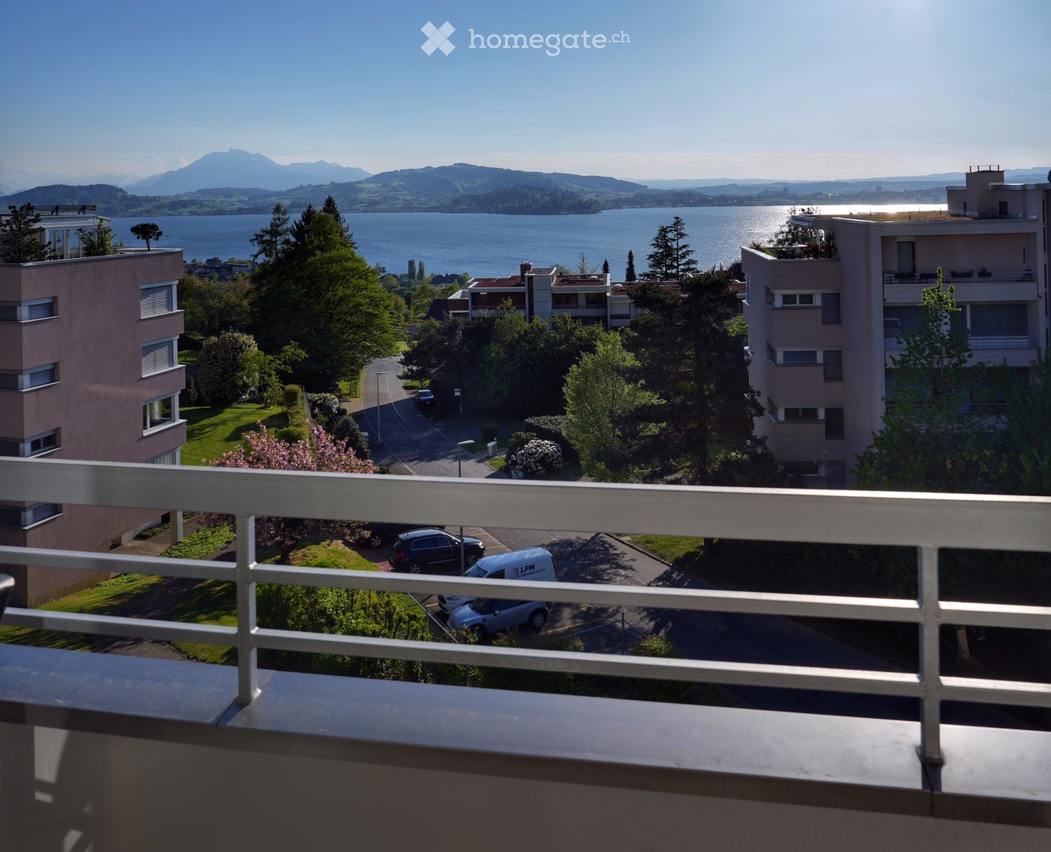 Balcony overlooking a residential area with parked cars, greenery, and a scenic lake and mountain view in the distance.