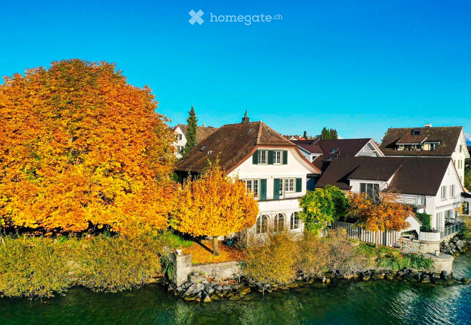 A picturesque residential area with traditional houses, surrounded by autumn-colored trees and a calm river.