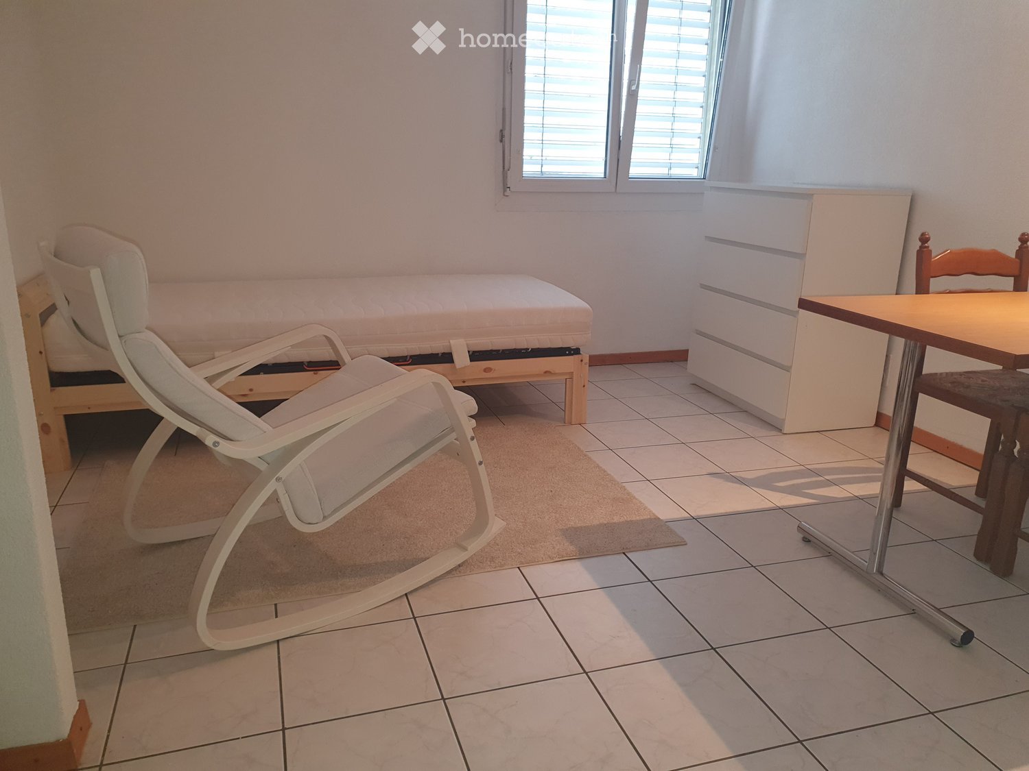 A furnished room with a bed, a white lounge chair, a small table, and a rug on the floor.