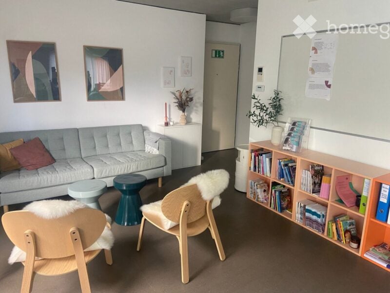 A modern, well-lit living room with a light grey sofa, colorful cushions, a small green stool, wooden chairs with white fur throws, a bookshelf filled with books, and decorative plants. The walls are white with several framed artworks.