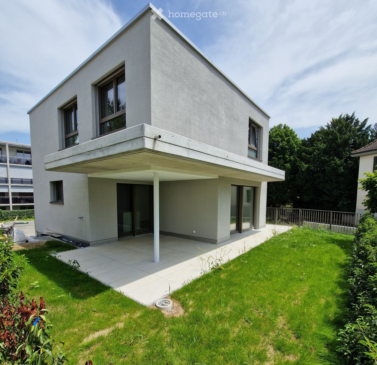 Modern two-story house with a flat roof, large windows, and a balcony on the upper floor. The house has a raised ground floor and is surrounded by a well-maintained lawn and garden.