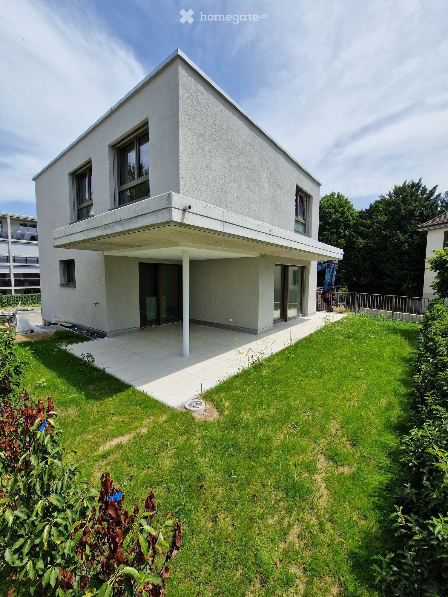 Modern two-story house with a flat roof, large windows, and a raised ground floor. The house features a spacious balcony and is surrounded by a well-maintained lawn and greenery.