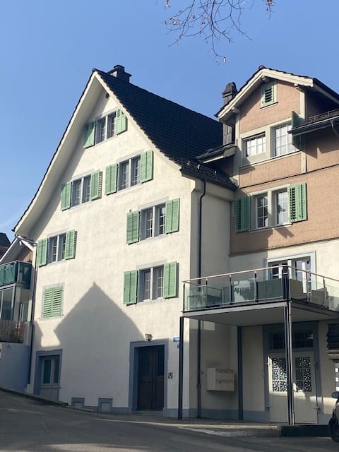 Two-story residential building with green shutters, white walls, and a balcony on the right side.