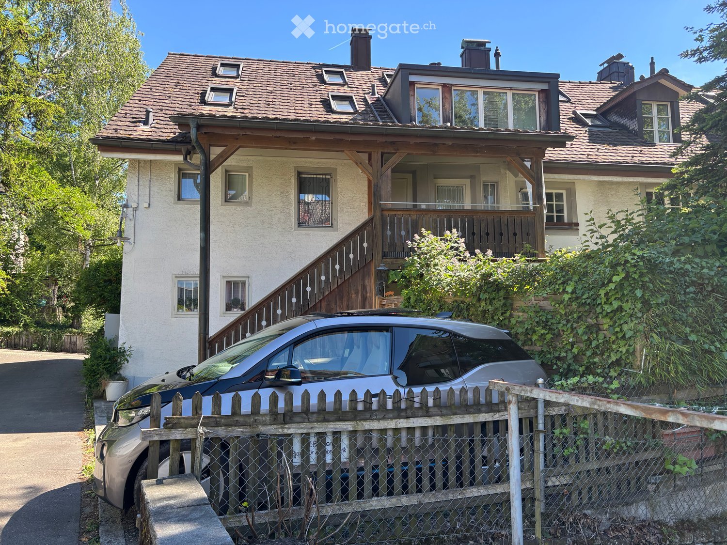 Two-story house with a tiled roof, multiple windows, and a wooden staircase leading to a balcony. A car is parked in front of a wooden fence.