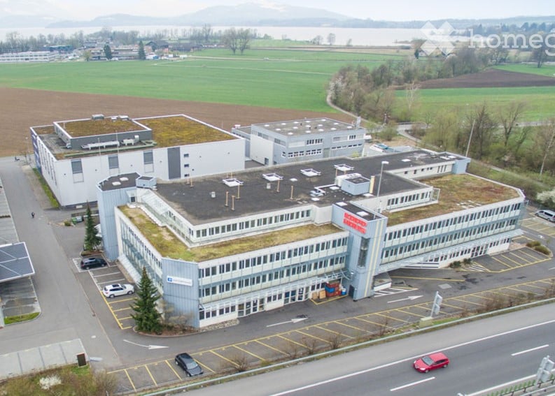 Modern industrial building with green rooftop, multiple sections, parking lot, and surrounding open fields.