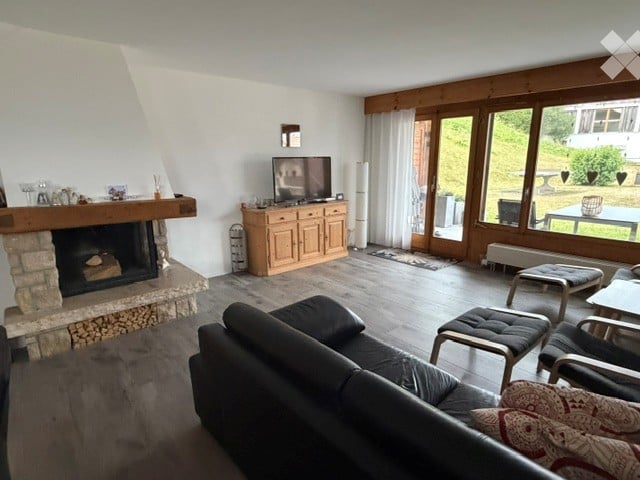 Modern living room with large windows offering a scenic view, featuring a black leather sofa, coffee table, fireplace, TV, and wooden cabinet.