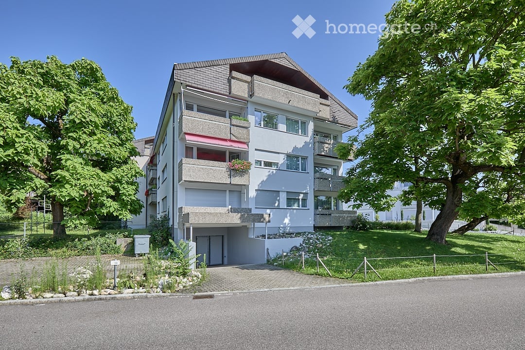 4-story white apartment building, multiple balconies, garden in front, paved driveway