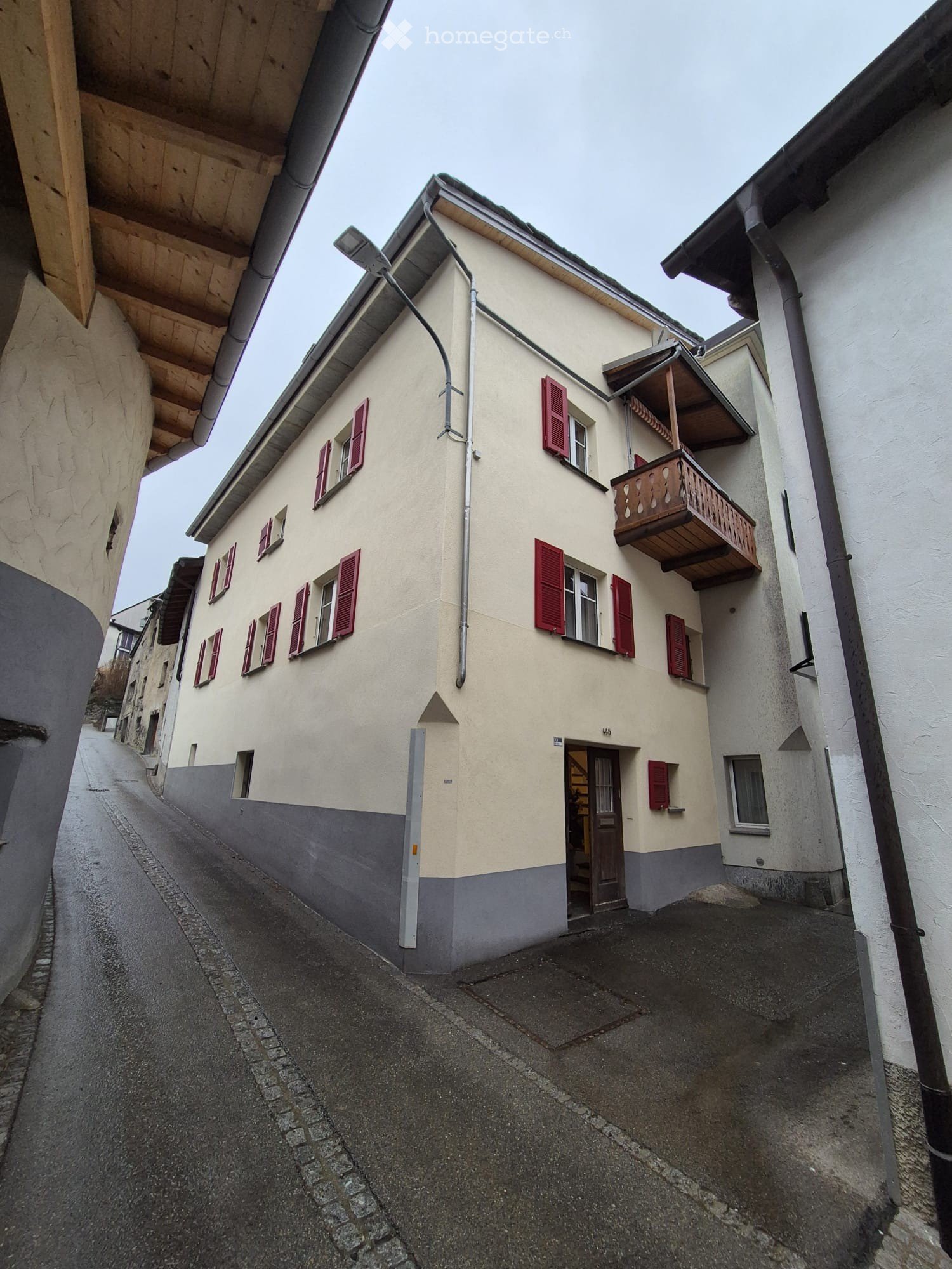 A two-story residential building with beige walls, red shutters, and a small balcony on the upper floor. The building is situated on a narrow, cobblestone street.