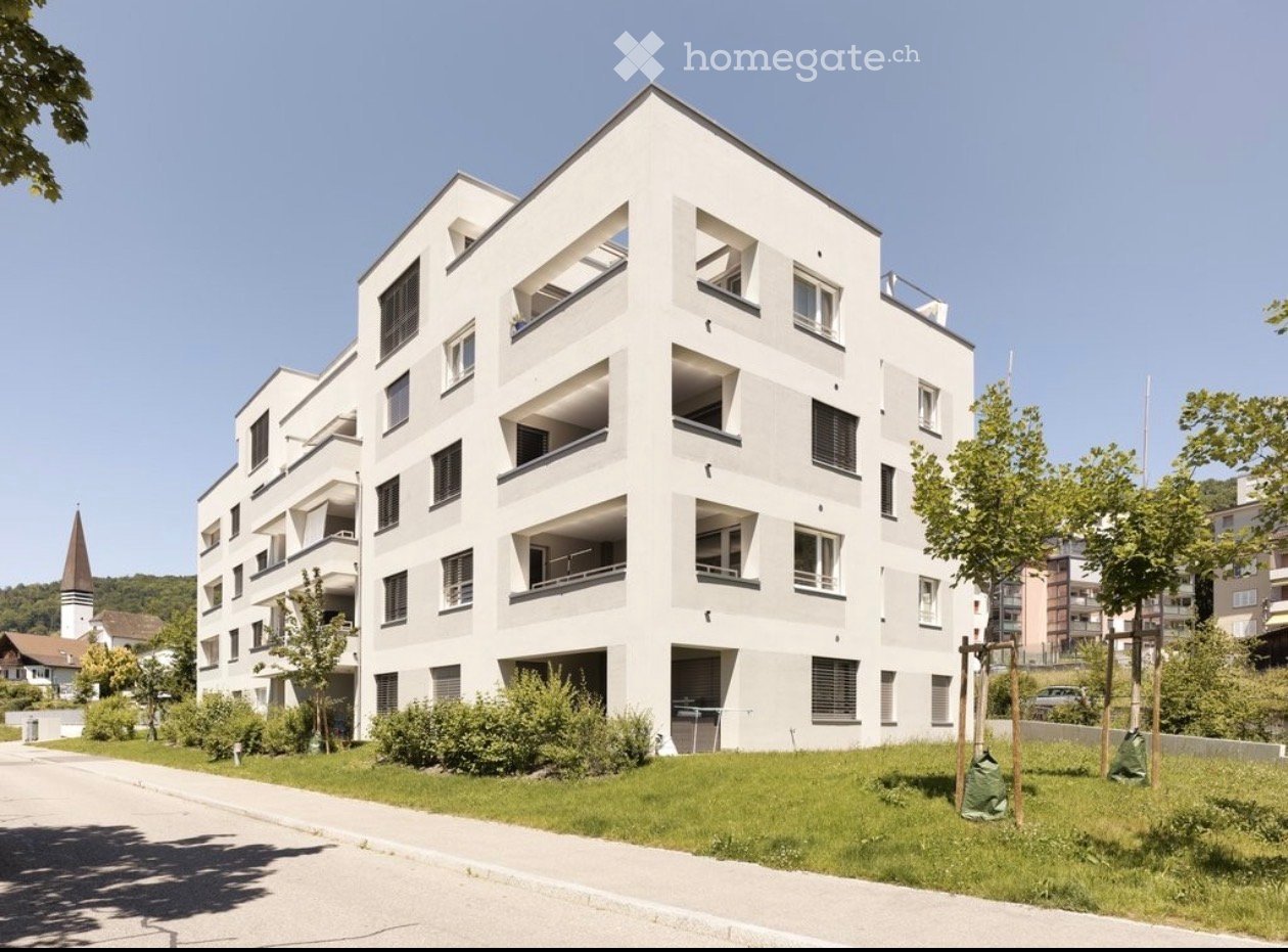 Modern multi-story residential building with white facade, numerous windows, and balconies. Surrounded by greenery and a paved pathway.