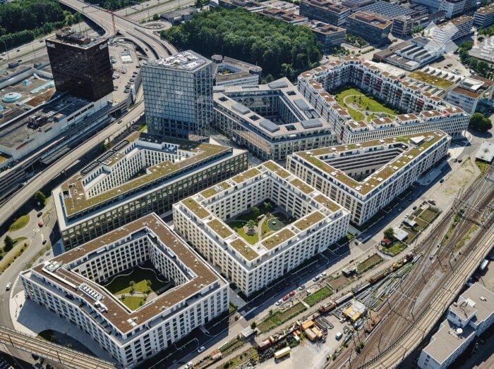 Aerial view of a modern urban development with multiple interconnected buildings featuring green roofs, surrounded by infrastructure and greenery.