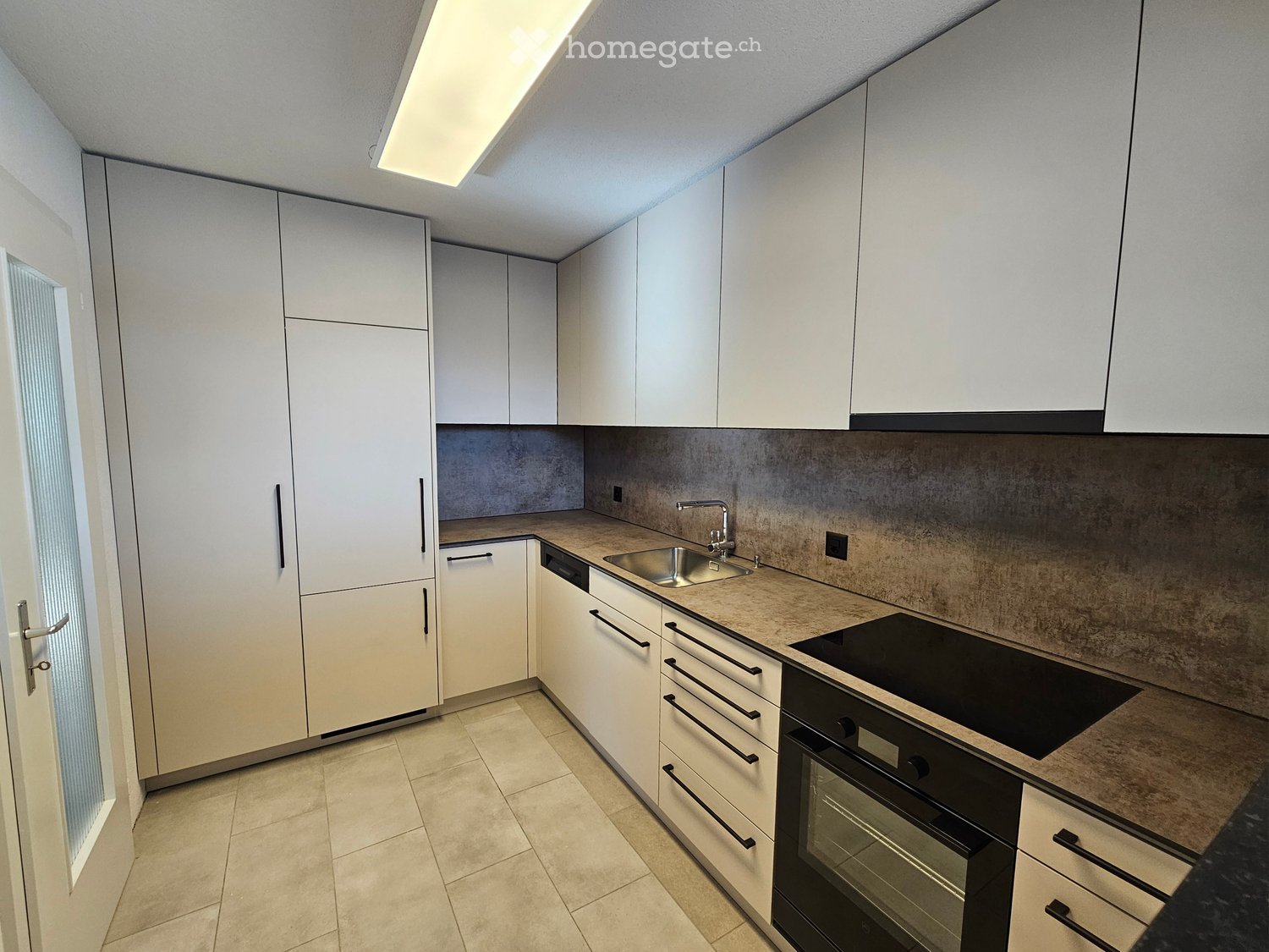 Modern kitchen with white cabinets, stainless steel sink, and built-in oven. The countertops are light brown, and the floor is tiled.