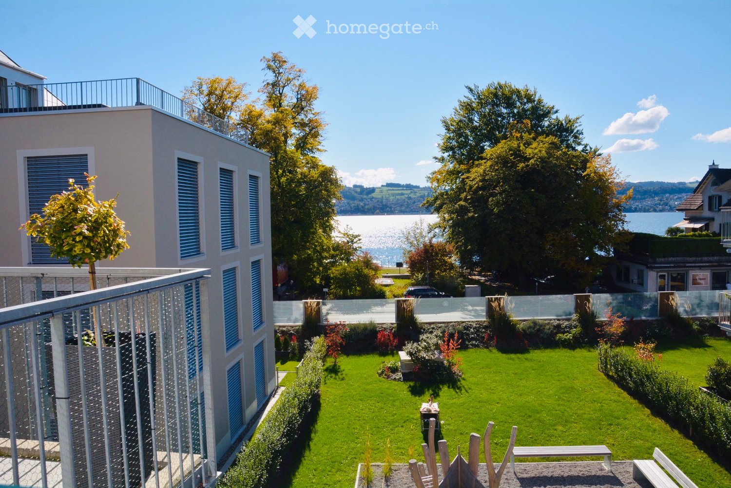 A well-maintained garden with a green lawn, small trees, shrubs, and a modern building with a balcony overlooking a scenic lake and distant mountains.
