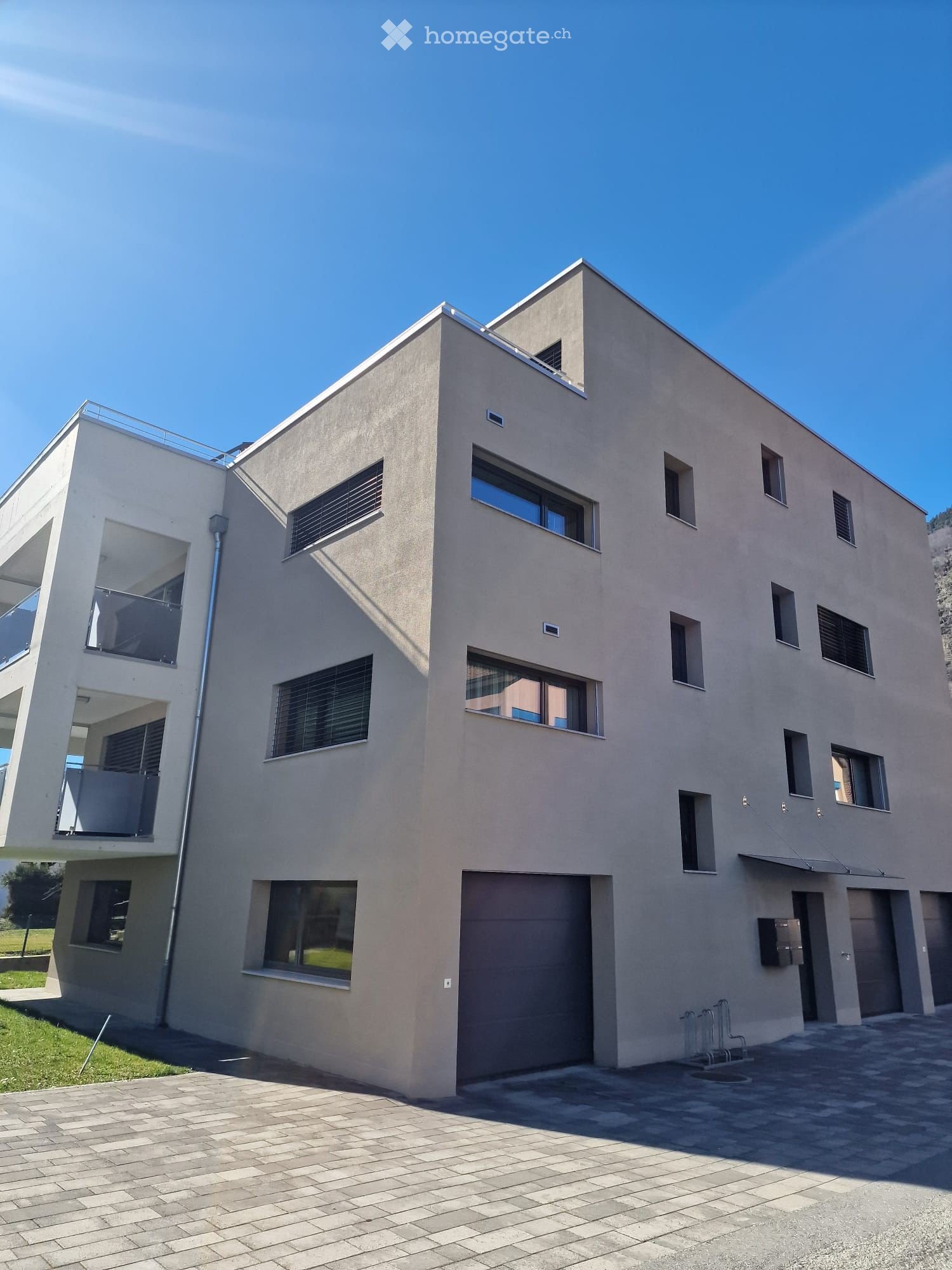 Modern two-story house with beige facade, large windows, and a garage entrance. Features a balcony on the upper level and a paved driveway.