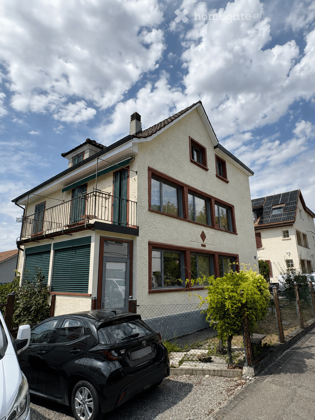 Two-story house with white facade, red window frames, balcony on the upper floor, and solar panels on the neighboring roof.