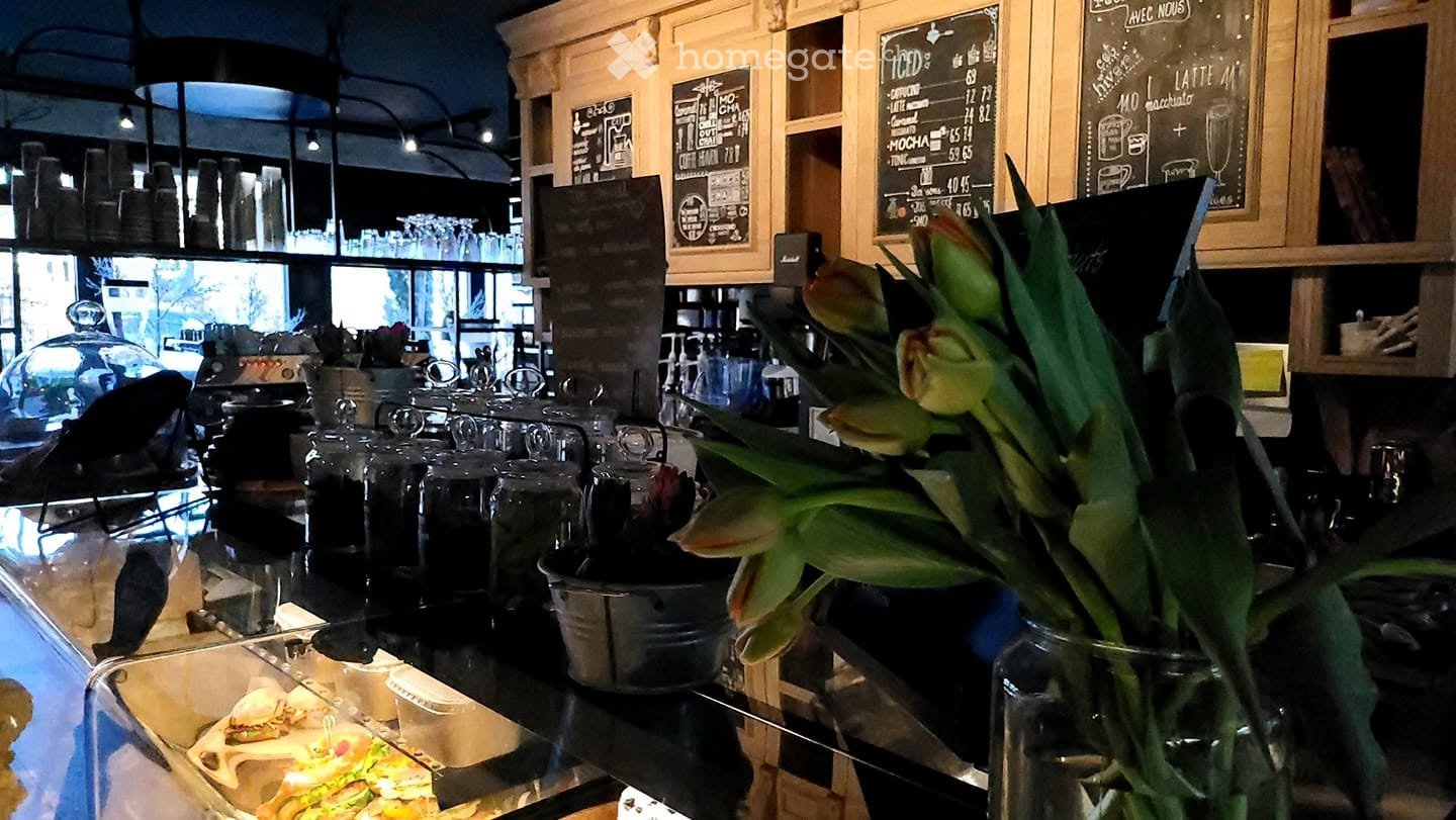 A cozy kitchen area with wooden cabinets, a menu board, a flower vase, and a coffee machine. The counter displays various cups and dishes.