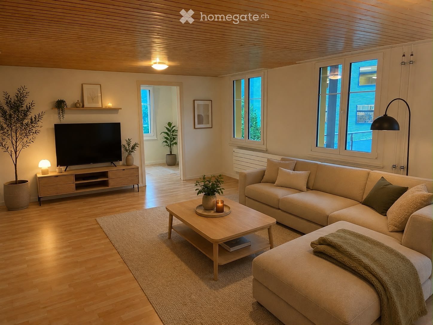 Modern living room with light wood flooring, white walls, and large windows. Features a beige sectional sofa with an ottoman, a wooden coffee table with a candle and books, a television on a wooden stand, and several potted plants. The room has a cozy, mi
