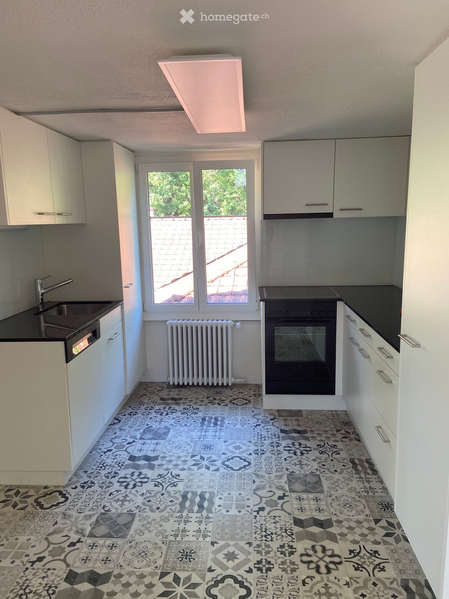 Modern kitchen with white cabinets, black countertops, a sink with a modern faucet, an oven, and a radiator. The floor features a decorative tile pattern in shades of grey and black.