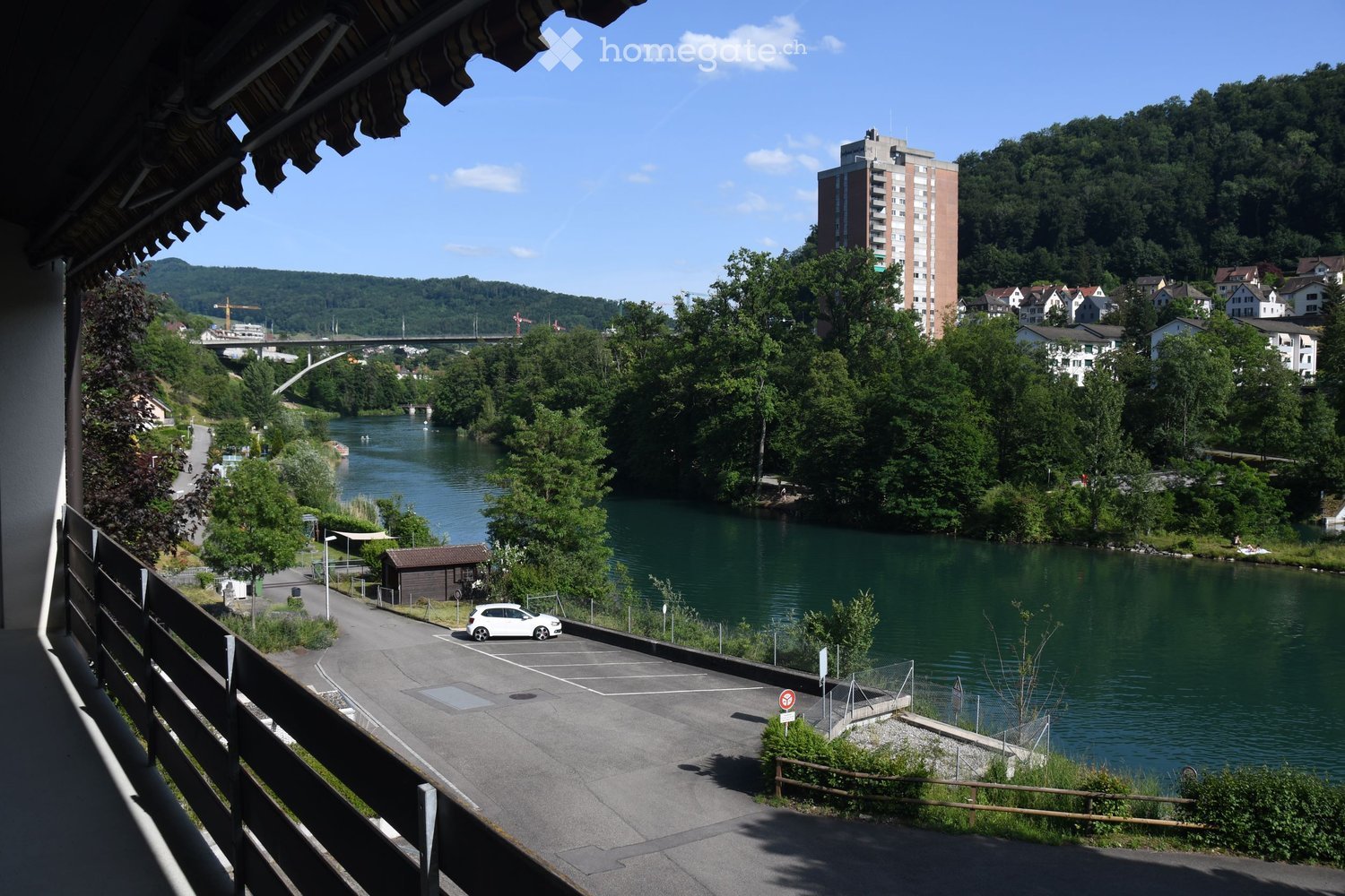 Balcony overlooking a river with a parking area, small shed, and distant buildings and hills.