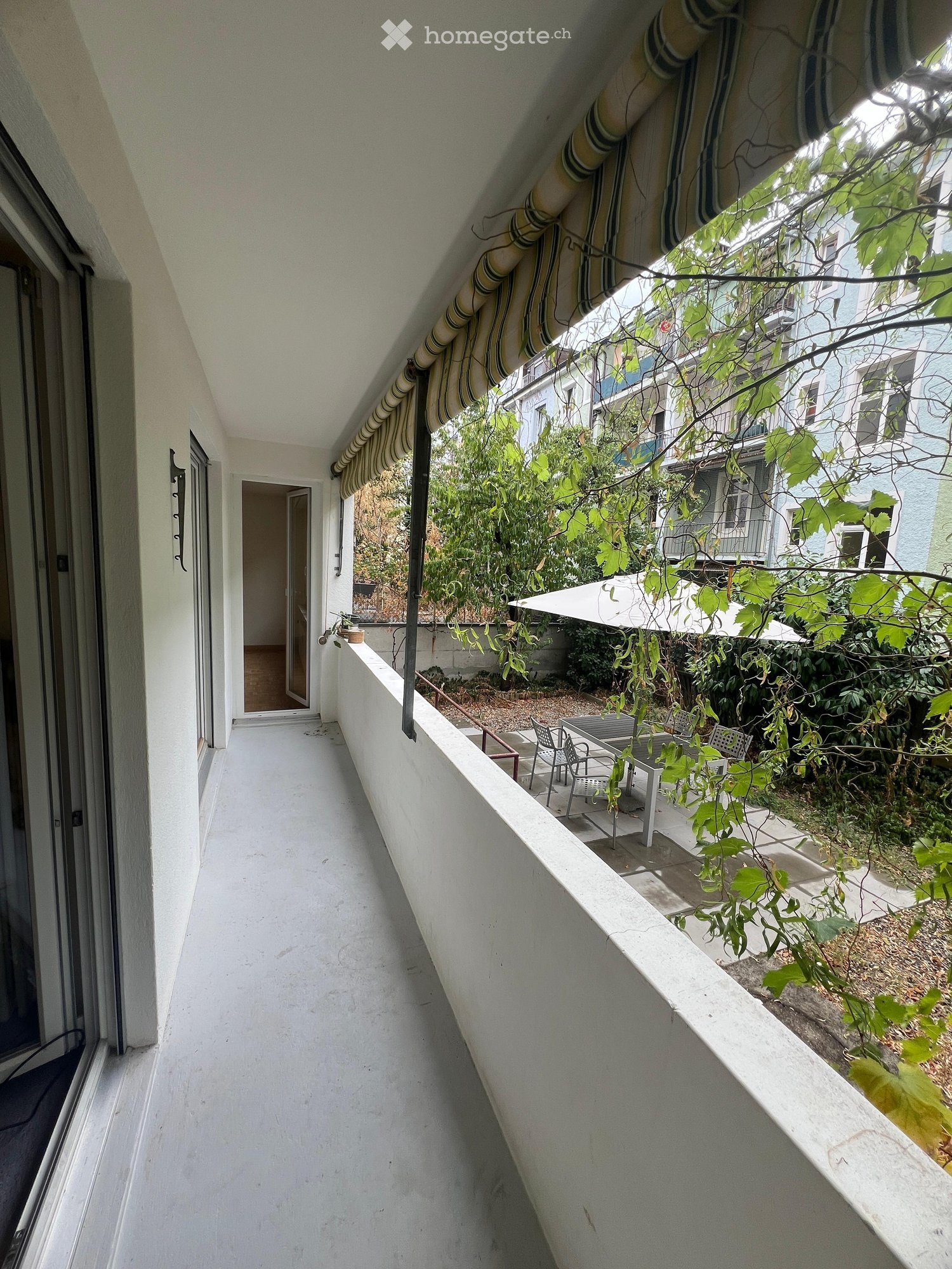 Balcony with white railing, striped awning, outdoor furniture, and greenery visible in the background.