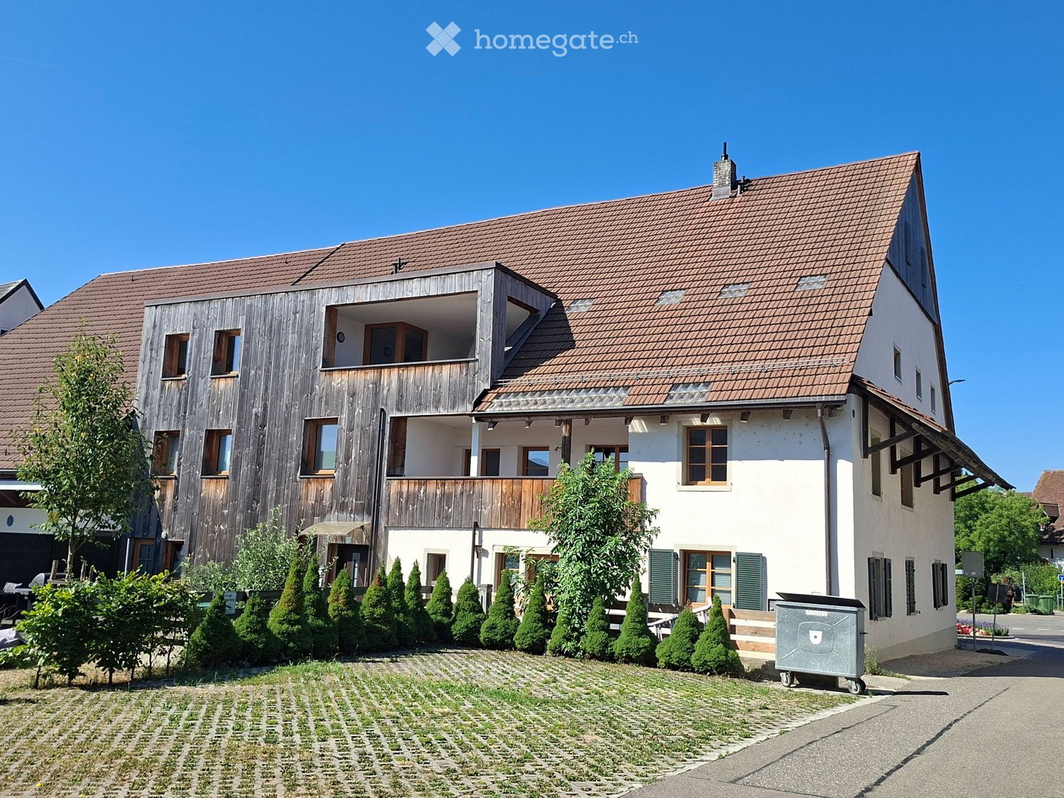 Two-story house with a tiled roof, wooden balcony, and well-maintained garden with trimmed shrubs and trees.